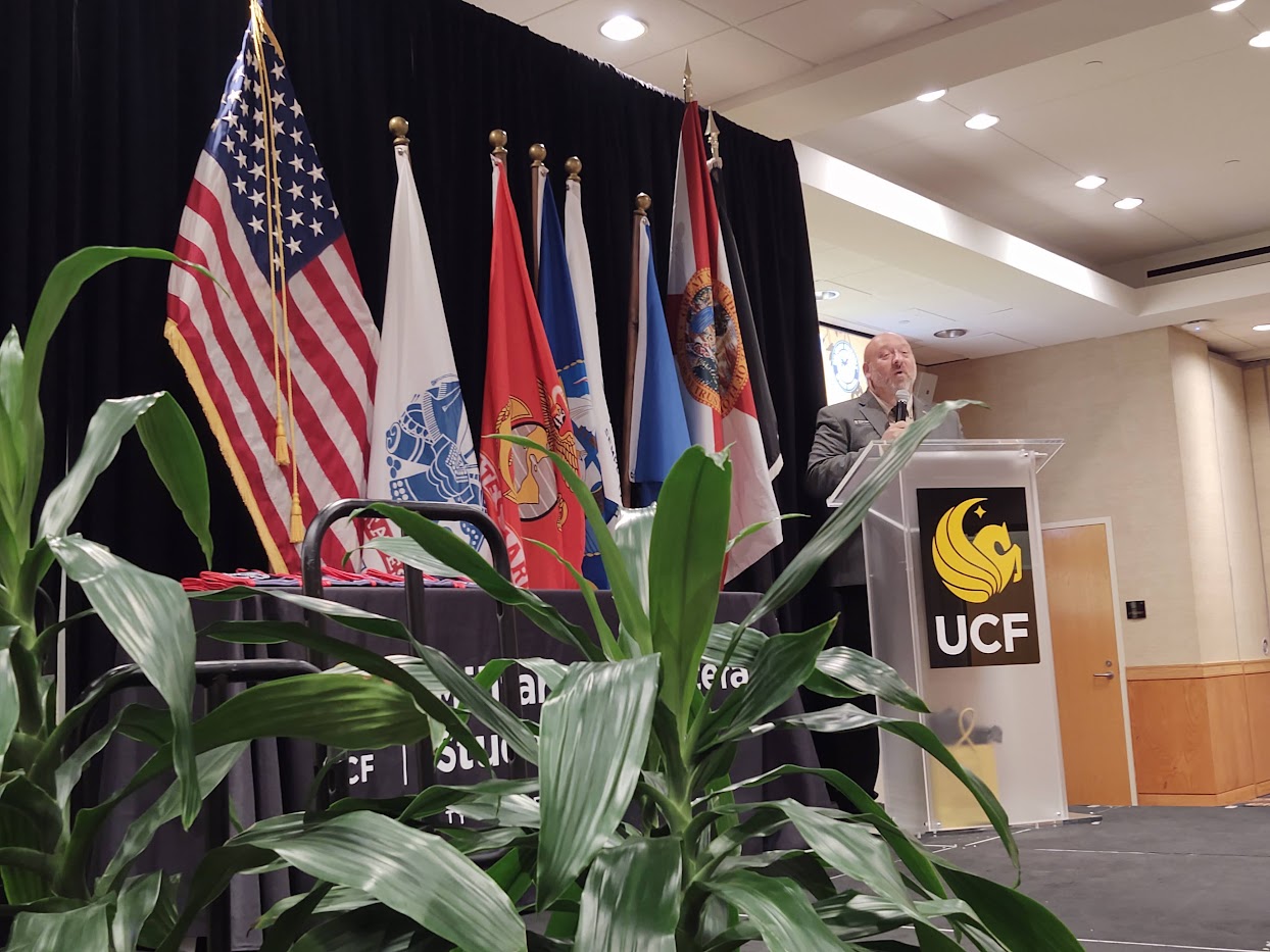 Michael Kepner speaking at a podium with the American Flag and other military flags behind