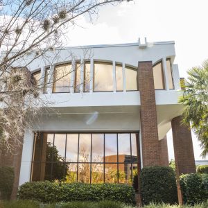 Millican Hall, a modern building at UCF with large windows surrounded by greenery, set against a bright sky.
