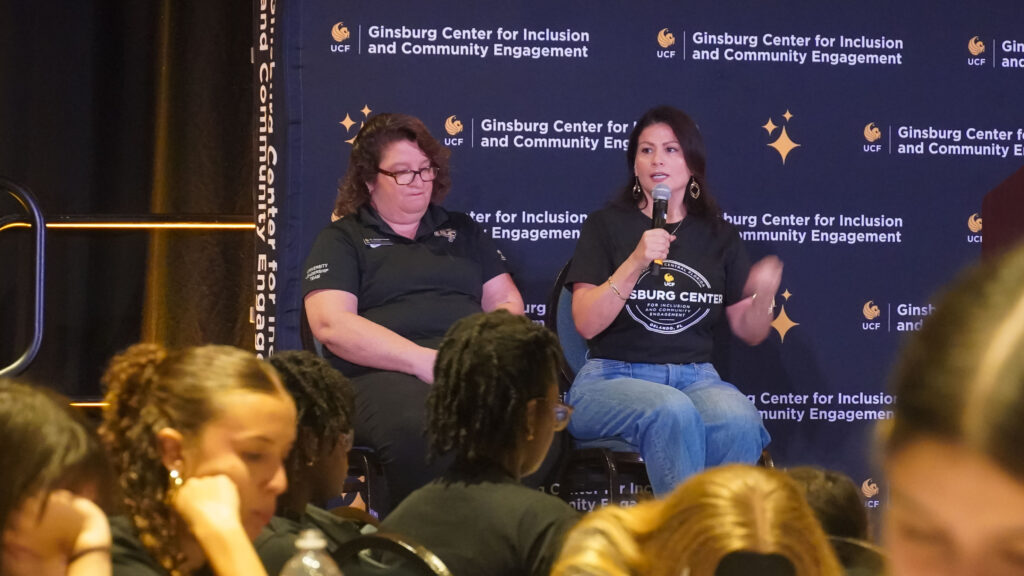 Two women participate in a panel discussion before an audience, with one speaking into a microphone and the Ginsburg Center for Inclusion and Community Engagement backdrop behind them.