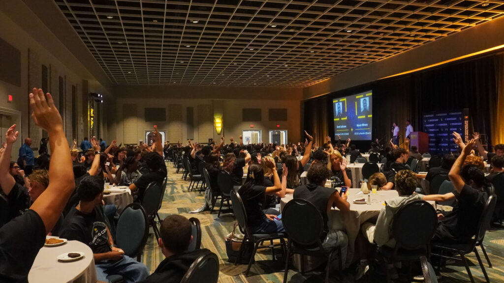Students raise their hands during a conference session in a ballroom while two speakers stand onstage.