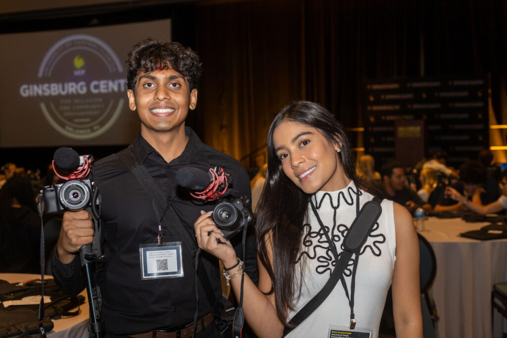 Two smiling event photographers pose with cameras in a dimly lit conference venue, with a “GINSBURG CENTER” projection behind them.