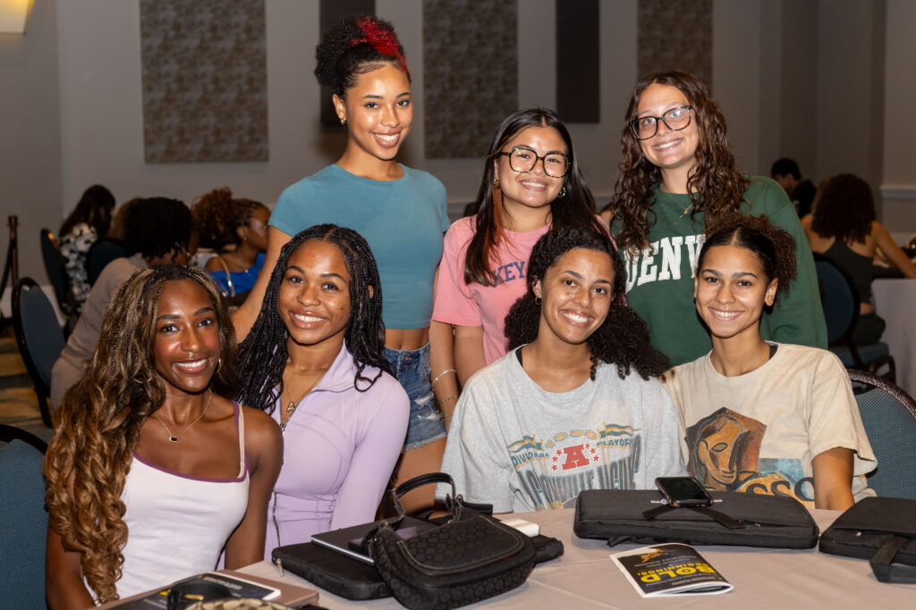 Seven smiling students pose together at a conference table, with other attendees and event materials visible in the background.