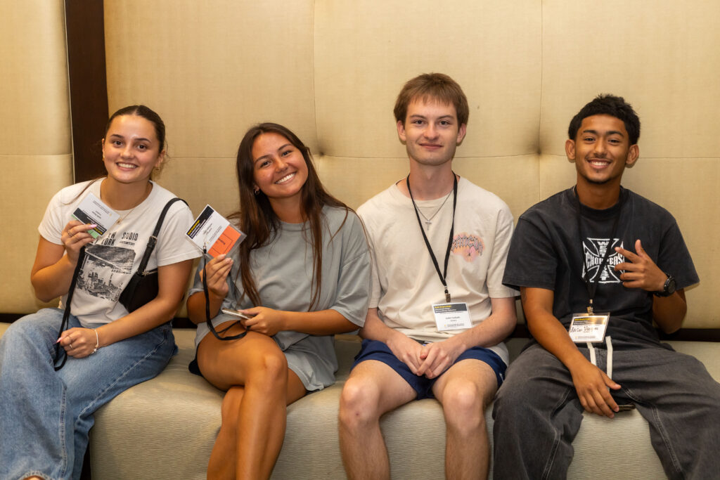 Four smiling students wearing conference badges sit together on a couch for a group photo.