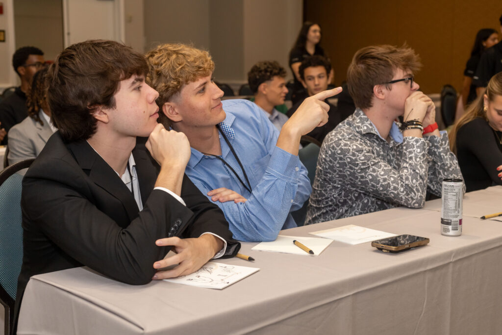 Students attend a conference, seated at a table with papers and pencils; one student points toward the front of the room.Students attend a conference, seated at a table with papers and pencils; one student points toward the front of the room.