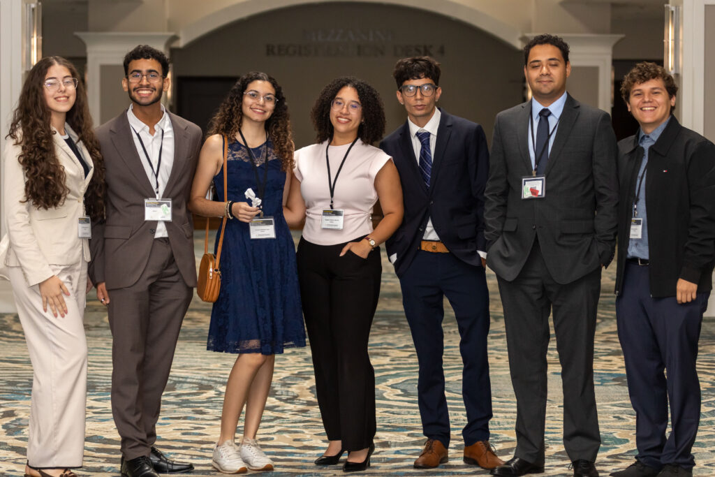 Seven event attendees pose together in formal and business attire in front of an indoor registration area.