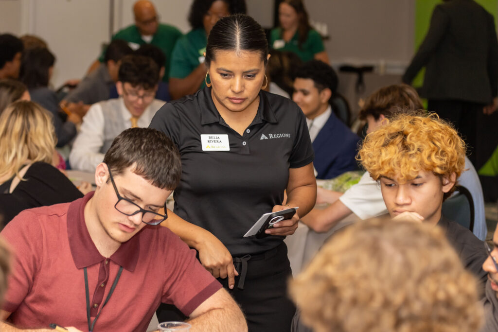Woman wearing a name badge stands among seated attendees at a student conference, holding a tablet or notebook.