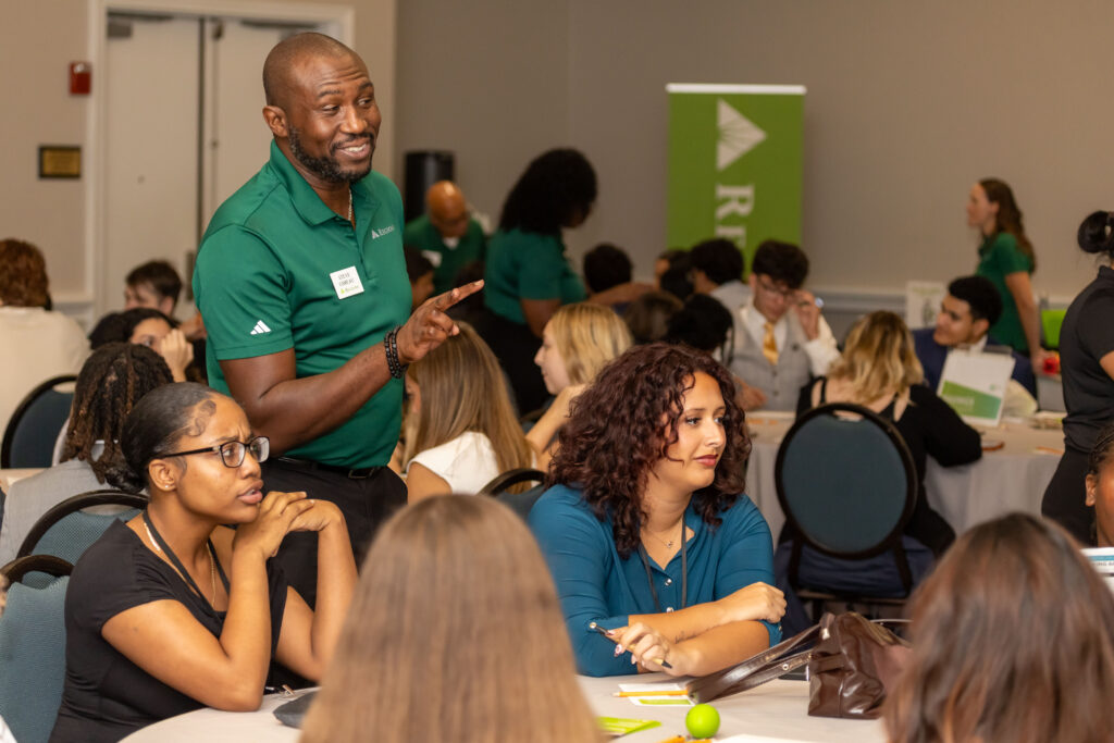 Attendees gather at a conference while a man in a green Regions polo speaks and gestures beside a table.