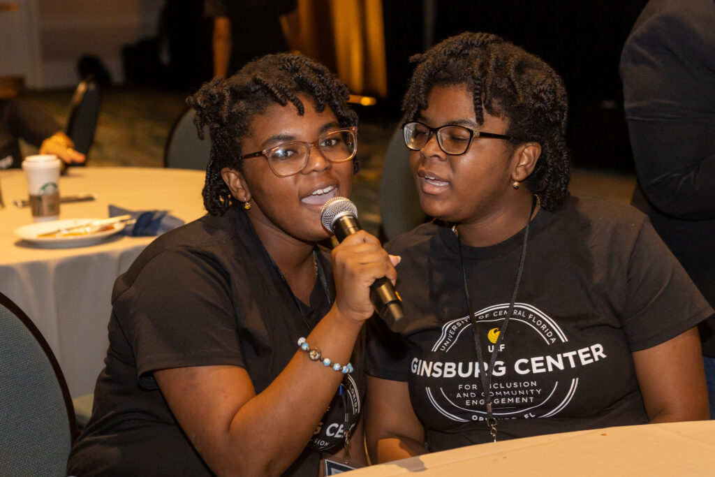 Two people sit together at an event, with one speaking into a handheld microphone while the other listens.