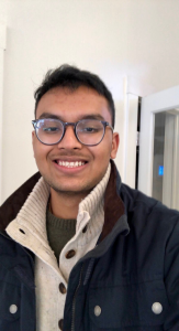 Headshot of Aayush Shah, wearing glasses, a dark jacket, and a scarf, smiling in an indoor setting with a white wall and doorway in the background.