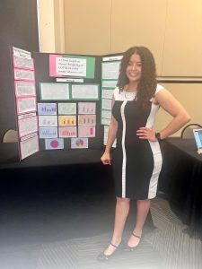 Dinamaris Aguilar standing confidently in front of a presentation board showcasing STEM-related materials, wearing a black and white dress, in a professional indoor setting.