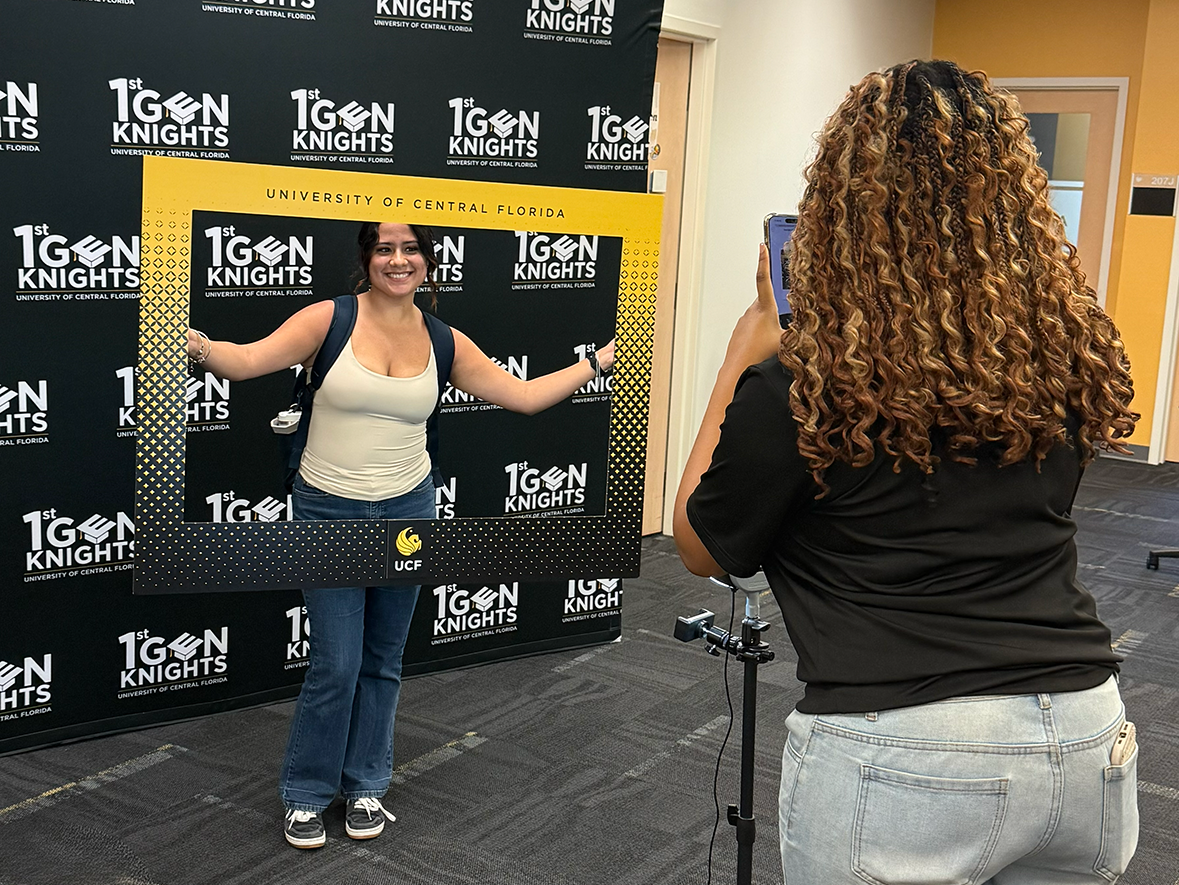 a student posing with a UCF themed frame around them and the First-gen knights backdrop behind while a staff member takes a photo