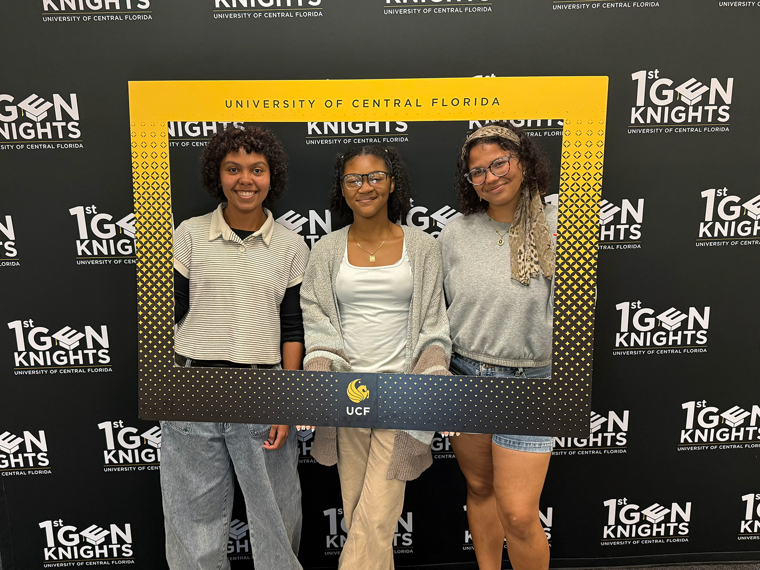 Three students posing with a UCF themed frame around them and the First-gen knights backdrop behind